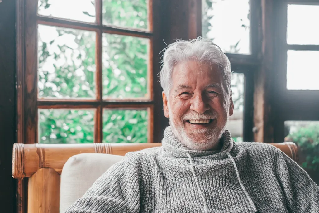 Older adult man sitting in a living room chair, smiling happily 